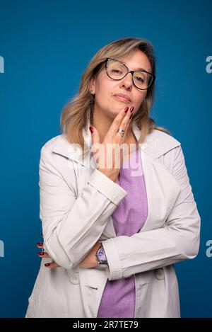 Portrait d'une femme médecin confiante vêtue d'un manteau beige, portant des lunettes, avec sa main sur son menton. Personne positive. Isolé sur blu Banque D'Images