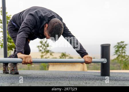 Jeune homme barbu habillé en entraînement noir sur une barbell sur le sol faisant des étirements dans une salle de sport en plein air lors d'une journée de pluie Banque D'Images