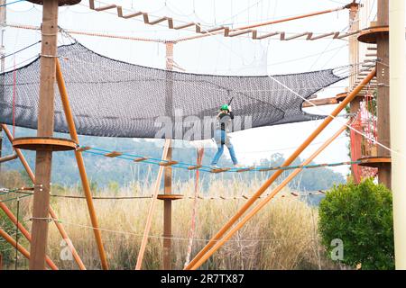 Adolescente dans l'équipement de harnais d'escalade, casque de sécurité vert de sport. Parc d'attractions de corde. Fixation fixant le mousqueton à la corde de sécurité. Hangin Banque D'Images