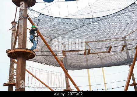 Adolescente dans l'équipement de harnais d'escalade, casque de sécurité vert de sport. Parc d'attractions de corde. Fixation fixant le mousqueton à la corde de sécurité. Hangin Banque D'Images