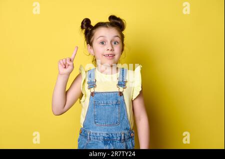 Studio portrait stupéfié petite fille de jeune pointe son index vers le haut, montrant une copie espace d'annonce sur fond jaune isolé Banque D'Images