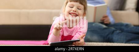 Petite fille de bébé souriant avec smartphone se trouve sur le sol en arrière-plan papa lit un livre Banque D'Images