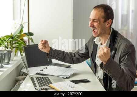 Concept de victoire. Joyeux et chanceux, un homme d'affaires caucasien barbu, pdg, gérant, est assis au bureau, en faisant des gestes avec des poings Banque D'Images