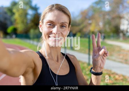 Athlète féminine après avoir fait du jogging dans le stade, discutant avec des amis et enregistrant un blog vidéo en ligne, blonde mature avec casque regardant la caméra de smartphone sourire, appel vidéo avec casque. Banque D'Images