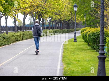 Un jeune homme conduit un scooter électrique, le long d'une allée de printemps à l'extérieur par temps clair. Mode de vie mobile. Vue de l'arrière. Copier l'espace. Banque D'Images