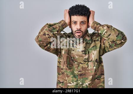 Homme arabe portant l'uniforme militaire de camouflage fou et effrayé avec les mains sur la tête, peur et surpris de choc avec la bouche ouverte Banque D'Images