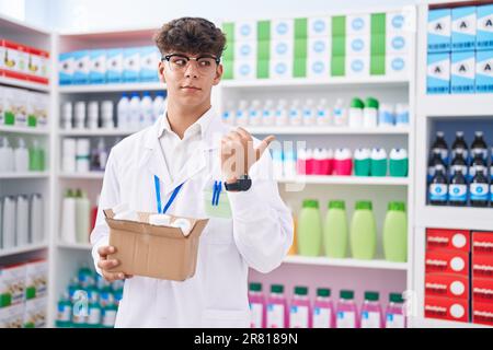 Adolescent hispanique travaillant à la pharmacie pharmacie drugstore boîte avec des pilules pointant le pouce vers le côté souriant heureux avec la bouche ouverte Banque D'Images