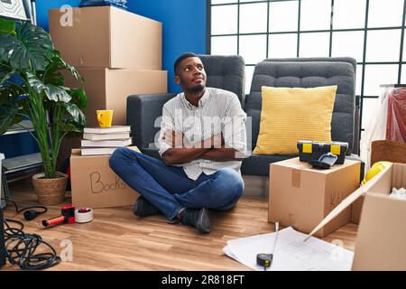 africain américain homme assis sur le sol dans une nouvelle maison regardant le côté avec les bras croisés convaincu et confiant Banque D'Images