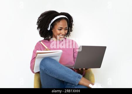 Joyeuse femme noire millénaire dans un casque sans fil assis dans un fauteuil, regarder la vidéo sur un ordinateur portable, prendre des notes Banque D'Images
