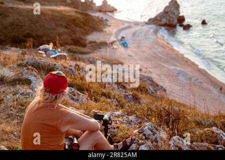 un homme s'assoit au sommet d'une montagne et tourne une vidéo sur une caméra au coucher du soleil sur la mer. Voyages et tourisme. Banque D'Images