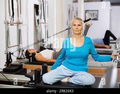 Une femme âgée vêtue de vêtements de sport est assise avec des jambes repliées sur l'équipement du reformeur et s'entraîne à nouveau dans le centre de réadaptation Banque D'Images