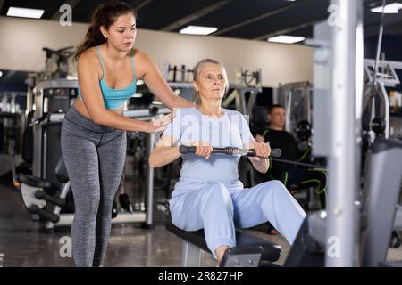 Une formatrice de forme physique féminine est engagée explique comment faire de l'exercice sur le simulateur à une femme âgée en salle de gym Banque D'Images