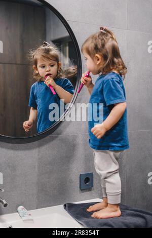 Petite fille caucasienne se brossant les dents dans la salle de bains regardant dans le miroir tout en utilisant la brosse à dents avec le dentifrice blanchissant. Heure du matin. Banque D'Images