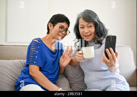Deux joyeuses femmes asiatiques d'âge moyen apprécient de parler avec leur petit-enfant par le biais d'un appel vidéo sur un smartphone tout en se relaxant dans le salon pour Banque D'Images
