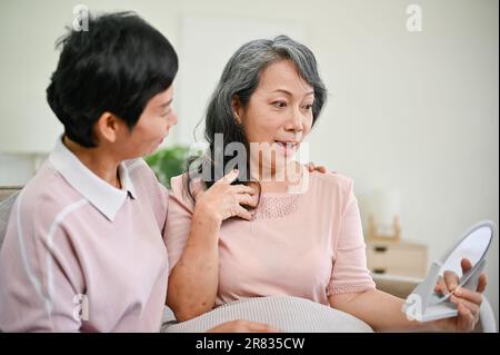 Une belle femme mûre d'âge moyen regarde son visage dans le miroir et se sent heureuse et choquée après avoir fait son maquillage avec son ami de hom Banque D'Images
