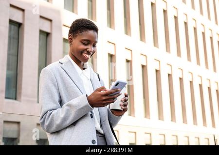 portrait horizontal d'une jeune femme africaine élégante. Elle se trouve près d'un immeuble de bureaux et utilise son smartphone Banque D'Images
