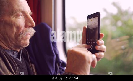 Homme âgé voyageant en train et prenant des photos du paysage par la fenêtre à l'aide d'un smartphone Banque D'Images
