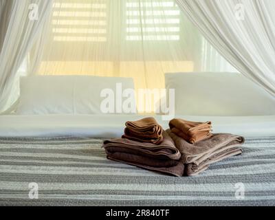 Ensemble de serviettes de bain brunes plies décorées sur le lit de matelas avec une couverture rayée et une moustiquaire blanche près de la fenêtre en bois dans la chambre à coucher de l'hôtel, asiatique s. Banque D'Images