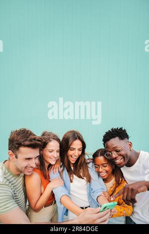 Portrait vertical d'un groupe multiracial de jeunes amis souriant à l'aide de leur application pour téléphone mobile sur fond bleu sarcelle. Divers étudiants adolescents qui s'amusent à regarder du contenu sur un réseau social. Photo de haute qualité Banque D'Images