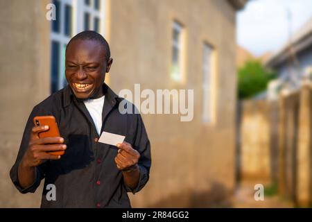 Homme africain excité portant une chemise noire avec son smartphone et une carte. transaction sans espèces Banque D'Images