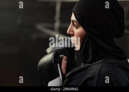 Gros plan de la jeune sportswoman en hijab noir, vêtements d'activité et gants de boxe debout devant le sac de poinçonnage et le regardant avant de frapper Banque D'Images