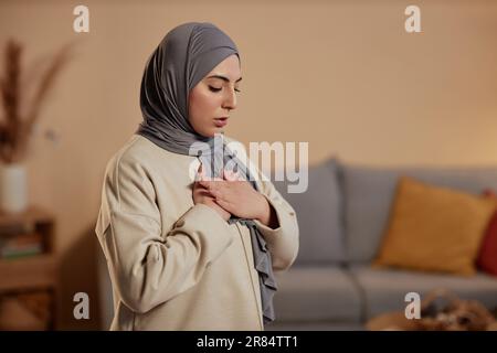 Jeune femme islamique en hijab gris et sweatshirt beige exprimant sa gratitude à son Dieu pendant la prière tout en gardant ses mains sur la poitrine Banque D'Images