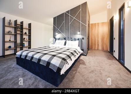 Chambre meublée simplement avec grand lit et étagères en bois avec décoration bouddhiste à la maison. Vue latérale d'un lit sombre recouvert d'un couvre-lit en tissu écossais sur le mur gris de la chambre. Concept d'intérieur. Banque D'Images