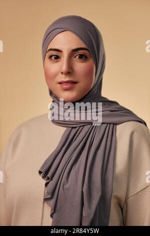 Jeune belle femme musulmane en foulard gris et pull beige regardant l'appareil photo tout en se tenant en isolement sur fond pastel Banque D'Images