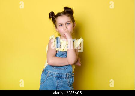 Adorable petite fille d'enfant debout avec les bras pliés, regardant pensif, isolé sur fond jaune de couleur. Enfants intelligents Banque D'Images