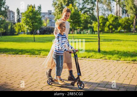 Mère s'amusant avec son fils dans le parc le jour ensoleillé. Le garçon est en scooter. Banque D'Images