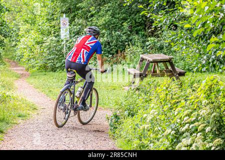 Vue arrière d'un homme senior portant un t-shirt Union Jack, faisant du vélo sur son vélo à travers un parc rural britannique en été. Banque D'Images