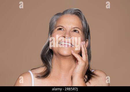 Une femme européenne aînée souriante aux cheveux gris touche le visage avec la main, regarde l'espace vide Banque D'Images