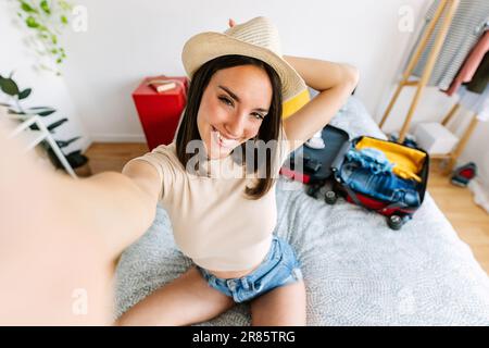 Jeune femme prenant le selfie tout en préparant la valise de vacances d'été à la maison Banque D'Images