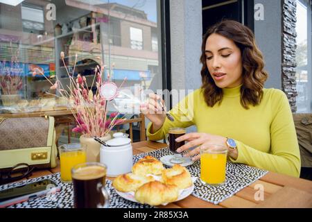 jeune femme latine argentine, assise à l'extérieur de la cafétéria, prenant le petit déjeuner avec du sucre dans le café, avec des croissants et du jus d'orange sur la ta Banque D'Images