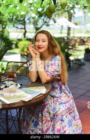 Portrait d'une belle femme blonde artiste et illustrateur assis à la table avec des dessins, des pinceaux et de la peinture Banque D'Images