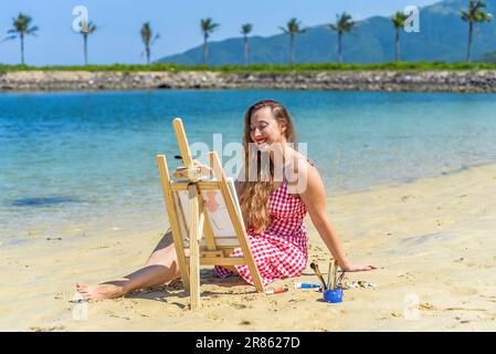 Jeune artiste femme assise à la plage avec un pot avec des pinceaux et un chevalet près de la mer et des palmiers en été Banque D'Images