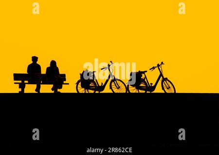 Couple de cyclistes âgés se reposant sur un banc à côté de leurs deux vélos silhouettés contre le ciel jaune de coucher de soleil en été Banque D'Images