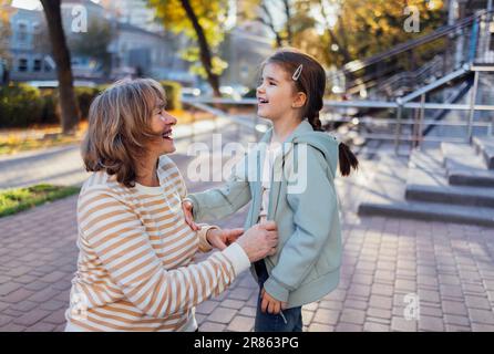 Femme âgée souriante vêtue de vêtements décontractés et petite-fille mignonne jouant dans la rue. Rire grand-mère d'âge moyen et mignon petit Banque D'Images