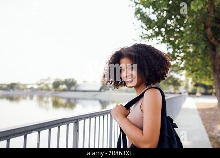 Jolie fille de course mixte tient sac à dos et sourit à l'appareil photo. Bonne mignonne fille d'école africaine mignonne avec des cheveux bouclés à la clôture sur la rive de la rivière. Beau f Banque D'Images