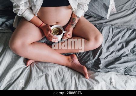 Jeune femme enceinte assise avec pattes croisées sur le lit. Tasse de thé photo dans la chambre en début de matinée. La santé de la femme, heureux prougn Banque D'Images