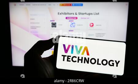 Personne tenant un téléphone portable avec logo de la conférence française Viva Technology (VivaTech) à l'écran en face de la page web d'affaires. Mise au point sur l'affichage du téléphone. Banque D'Images