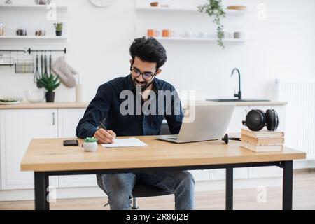 Beau indien portant une chemise sombre et des lunettes écrivant sur le document tout en restant à la maison pendant la mi-journée. Propriétaire d'entreprise efficace améliorant le contrat tout en vérifiant les données sur un ordinateur portable. Banque D'Images