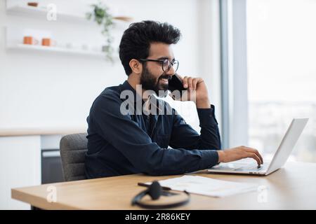 Vue latérale d'un homme d'affaires souriant qui tape sur un ordinateur portable tout en discutant sur un téléphone portable dans une pièce lumineuse par beau temps. Un responsable indien professionnel discute du rapport tout en regardant l'écran pendant l'appel. Banque D'Images