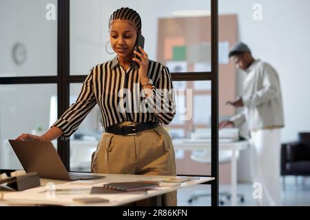 Jeune femme d'affaires confiante ou designer avec un smartphone près de l'oreille, debout près du lieu de travail avec un ordinateur portable et communiquant avec le client Banque D'Images