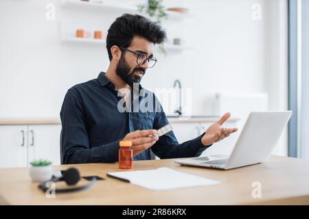 Adulte anxieux tenant des pilules rondes tout en ayant la conversation en ligne via ordinateur sur fond de cuisine. L'homme indien de la douleur recevant la consultation d'un médecin via une connexion Internet à la maison. Banque D'Images