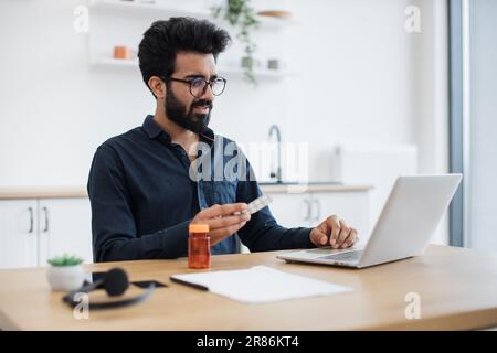 Adulte anxieux tenant des pilules rondes tout en ayant la conversation en ligne via ordinateur sur fond de cuisine. L'homme indien de la douleur recevant la consultation d'un médecin via une connexion Internet à la maison. Banque D'Images