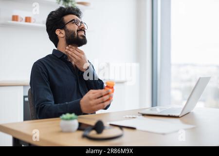 Jeune indien avec mal de gorge tenant des médicaments tout en parlant pendant la conversation en ligne avec le médecin de famille. Indépendant à distance efficace se sentant malsain après avoir attrapé la grippe à l'extérieur de la maison. Banque D'Images