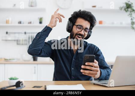 Joyeux homme en lunettes et chemise boutonnée en levant l'index tout en regardant le smartphone dans la cuisine. Joyeux adulte indien chantant tout en écoutant de la musique dans un casque pendant la pause. Banque D'Images