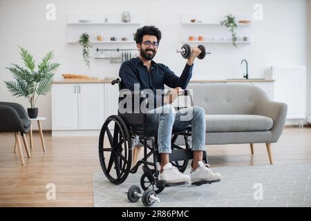 Vue en longueur d'un jeune homme indien avec mobilité réduite faisant des exercices assis avec poids dans la cuisine ouverte. Adulte positif en bapectakled dans les jeans gagnant des avantages de santé de l'entraînement. Banque D'Images