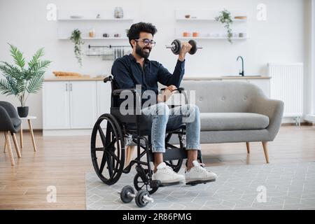 Vue en longueur d'un jeune homme indien avec mobilité réduite faisant des exercices assis avec poids dans la cuisine ouverte. Adulte positif en bapectakled dans les jeans gagnant des avantages de santé de l'entraînement. Banque D'Images